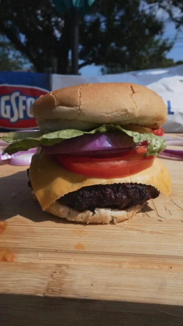 A hamburger stacked with cheese, tomato, onion and lettuce on a wood cutting board