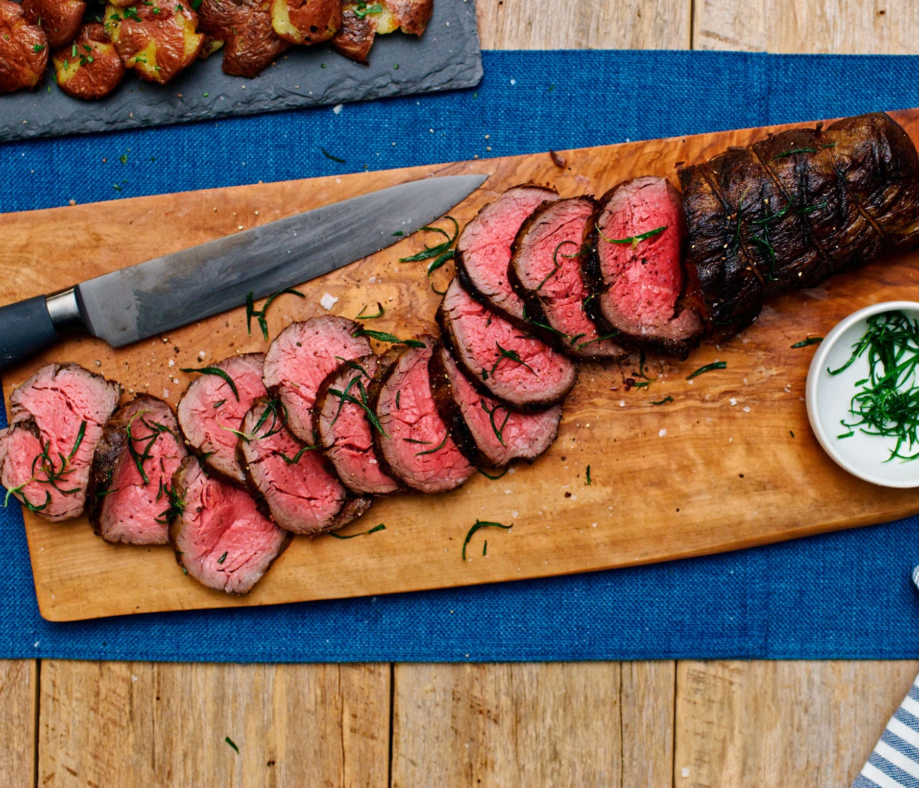 Sliced beef tenderloin with a seared crust arranged on a wooden cutting board beside fresh herbs and seasoning