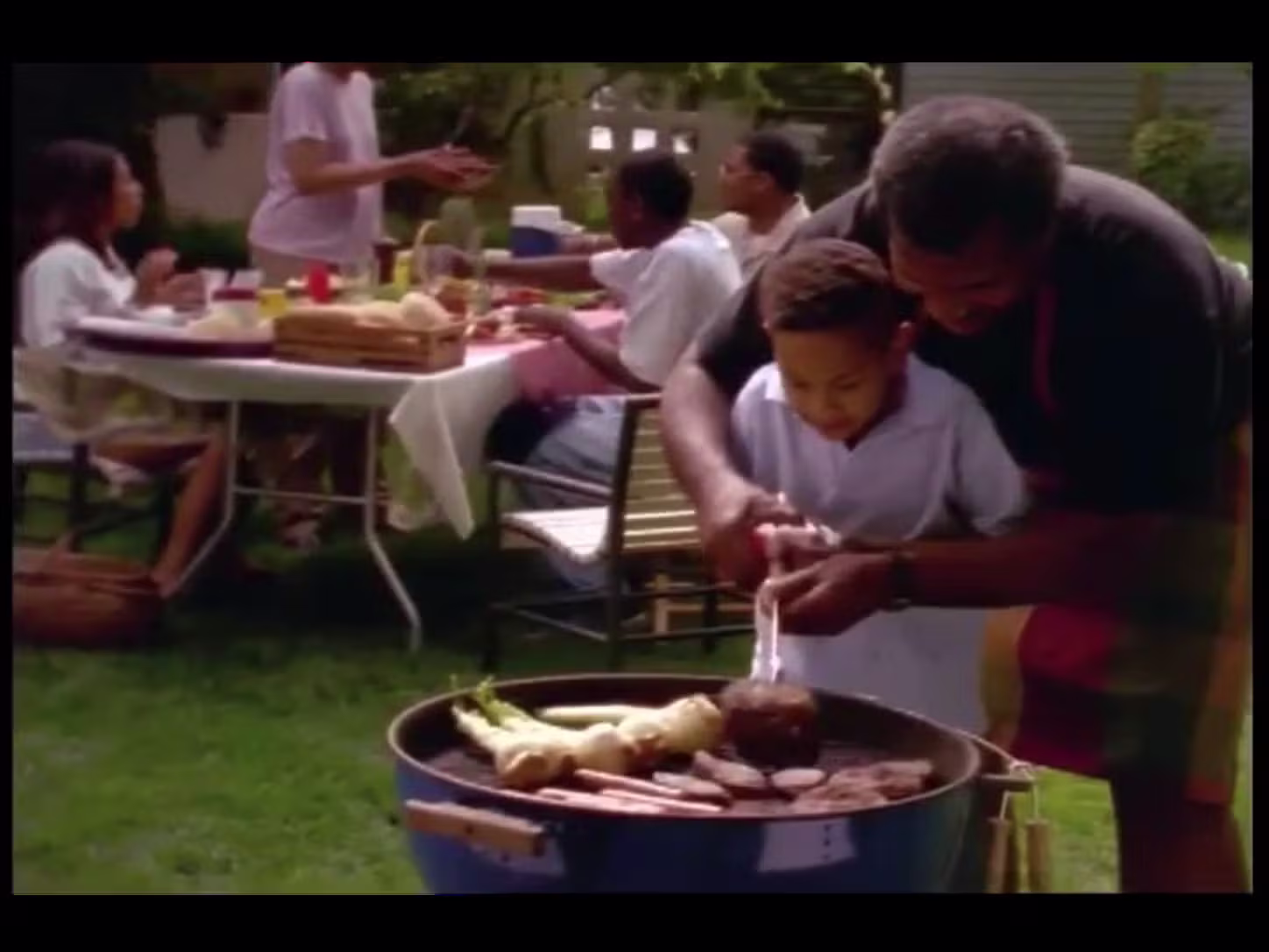 Man helping a child flip a hamburger on a grill. People at a picnic table in the background.