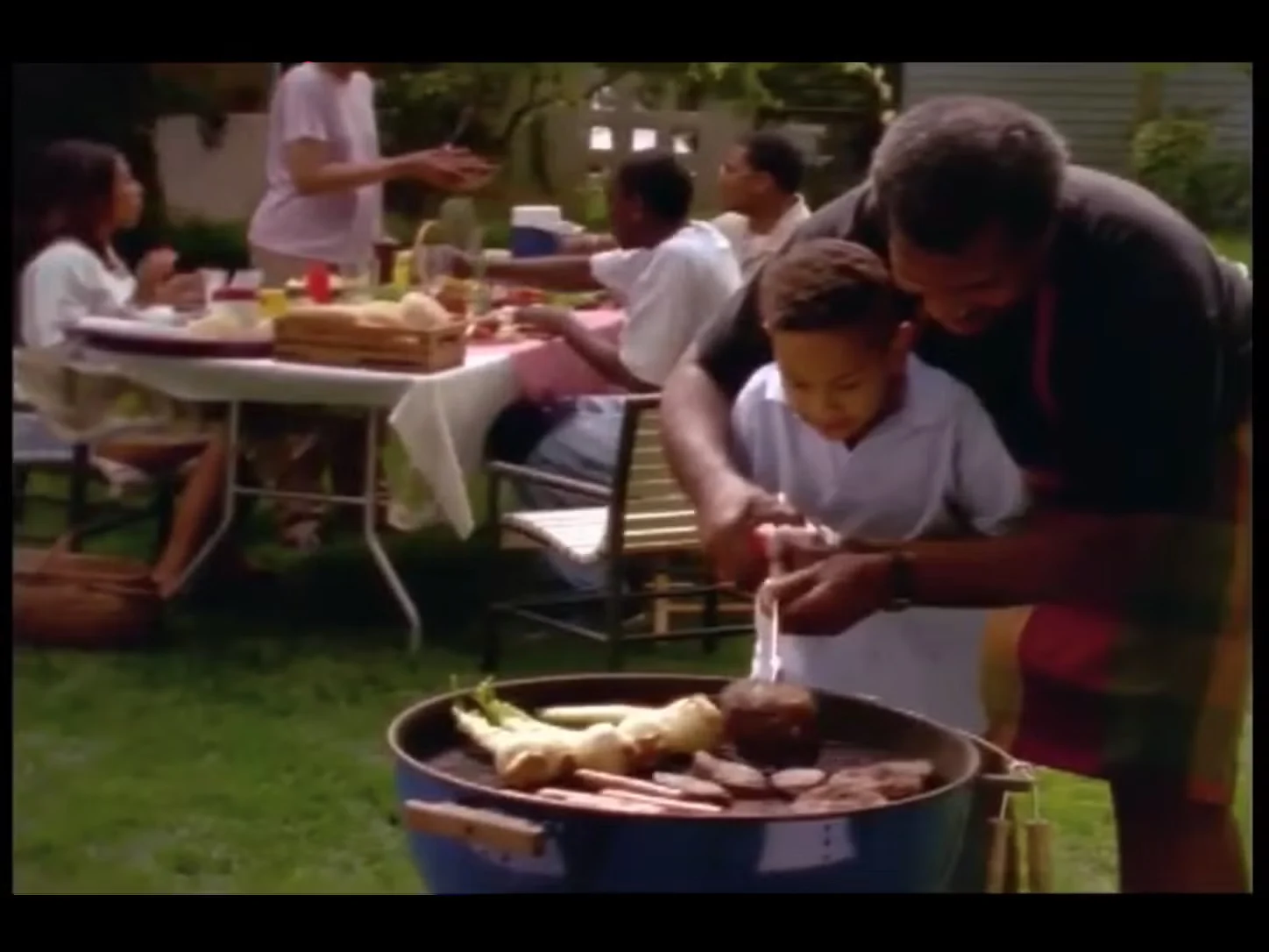 Man helping a child flip a hamburger on a grill. People at a picnic table in the background.