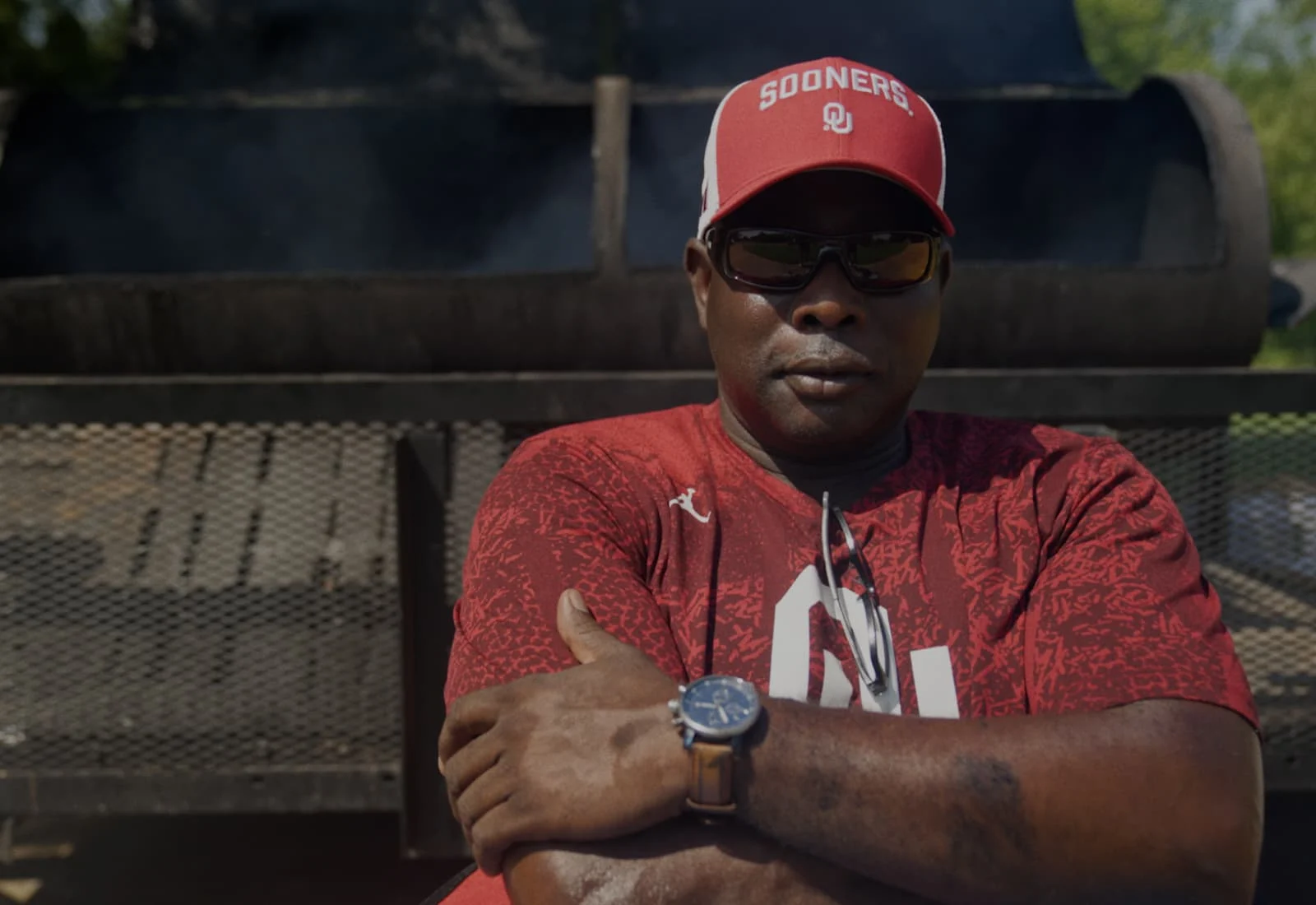 Rodney Dindy in University of Oklahoma hat and shirt standing in front of a grill with his arms folded