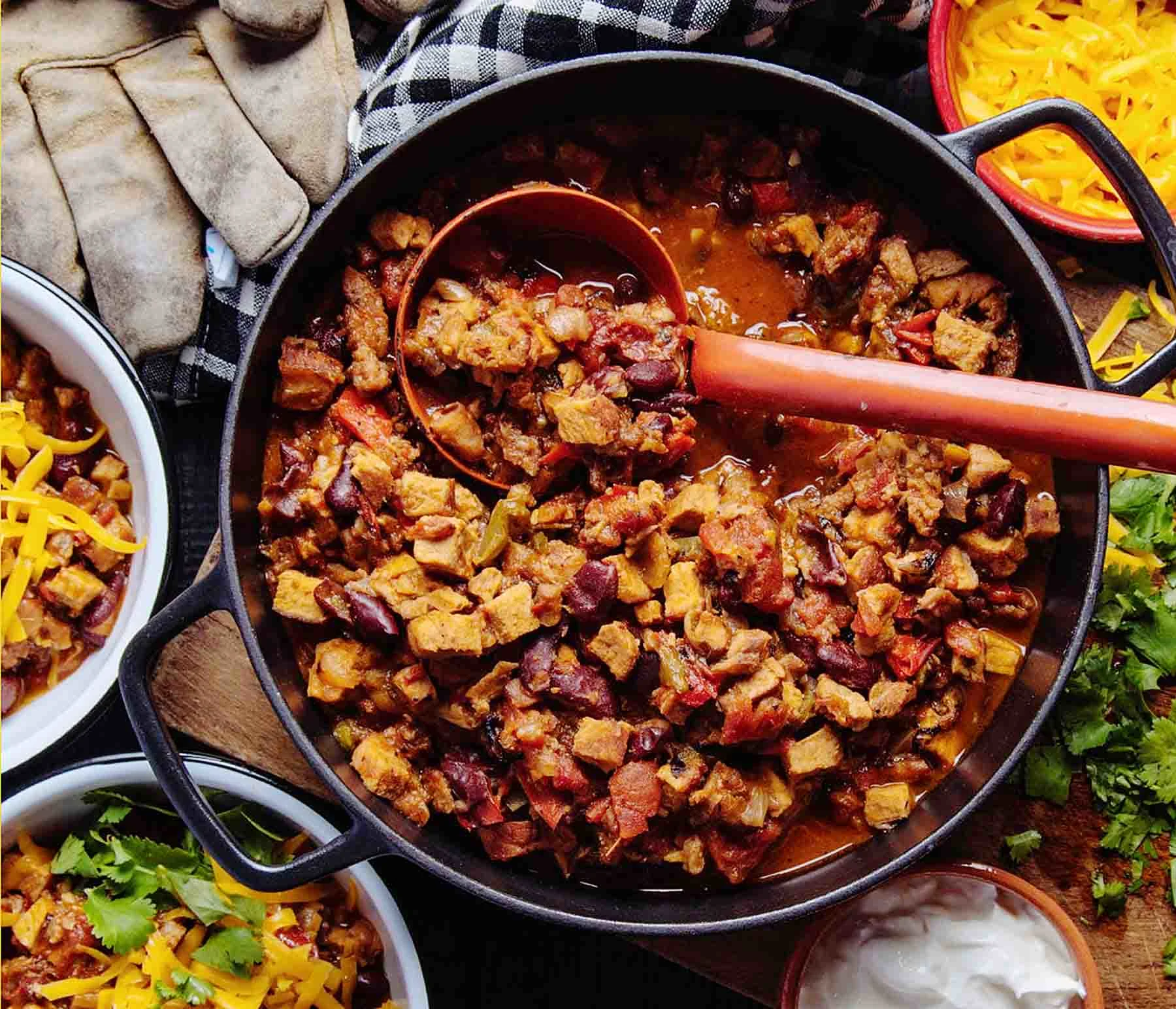 Rattlesnake chili in a cast iron pot with a red spoon, bowls of chili in the background