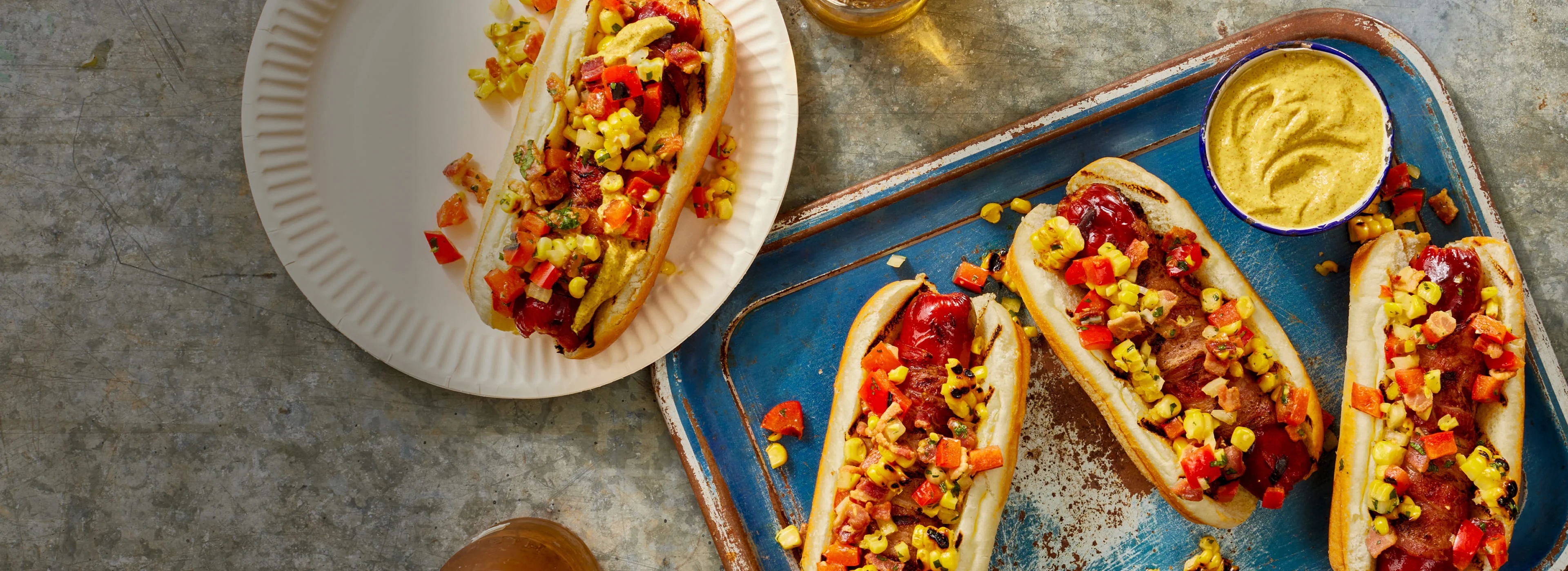Grilled hot dogs topped with corn salsa and bacon on a tray with mustard dipping sauce.