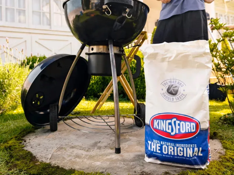 A person using a kettle grill with a bag of Kingsford charcoal in front