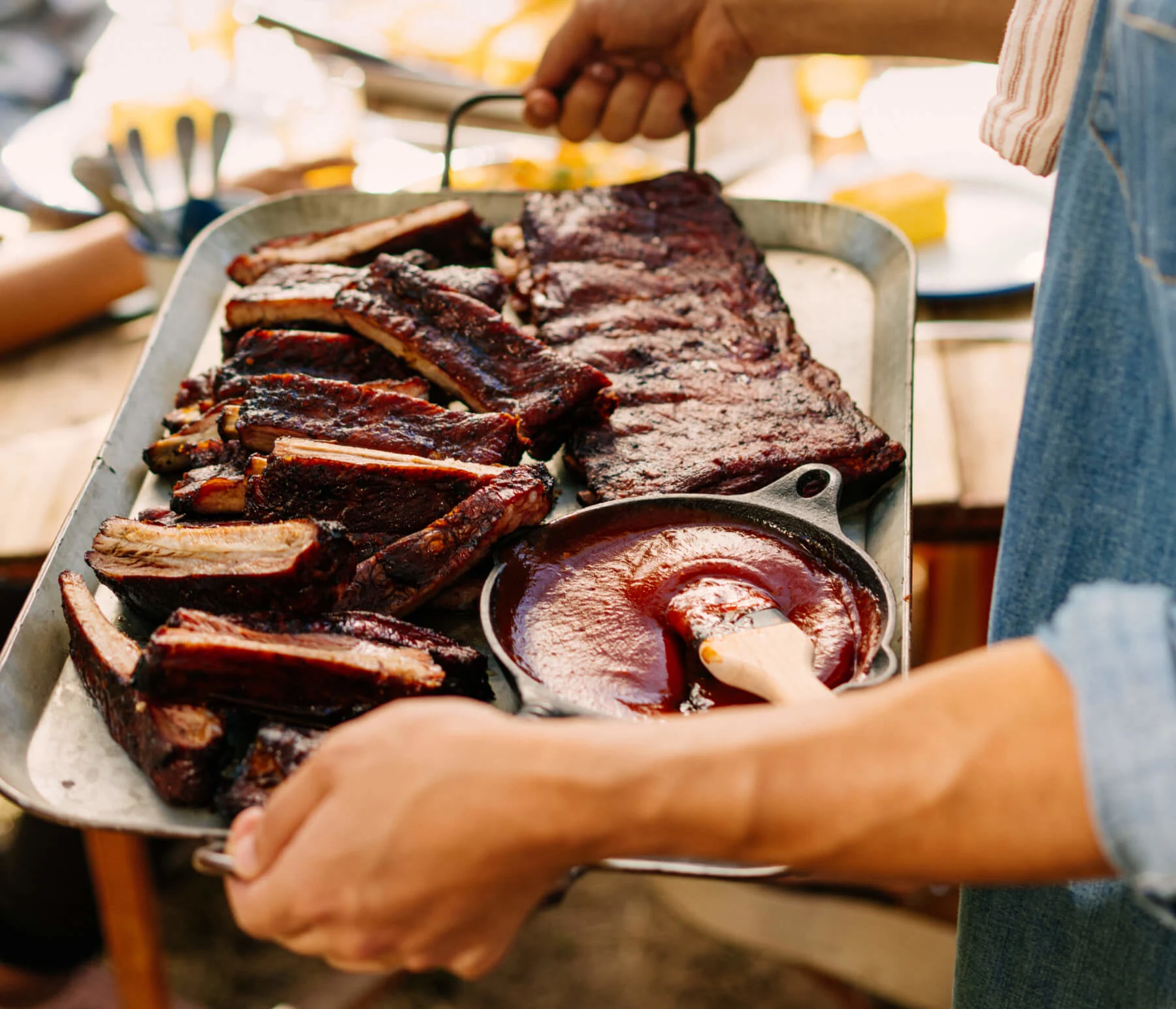 Person carrying a tray of grilled ribs and barbecue sauce