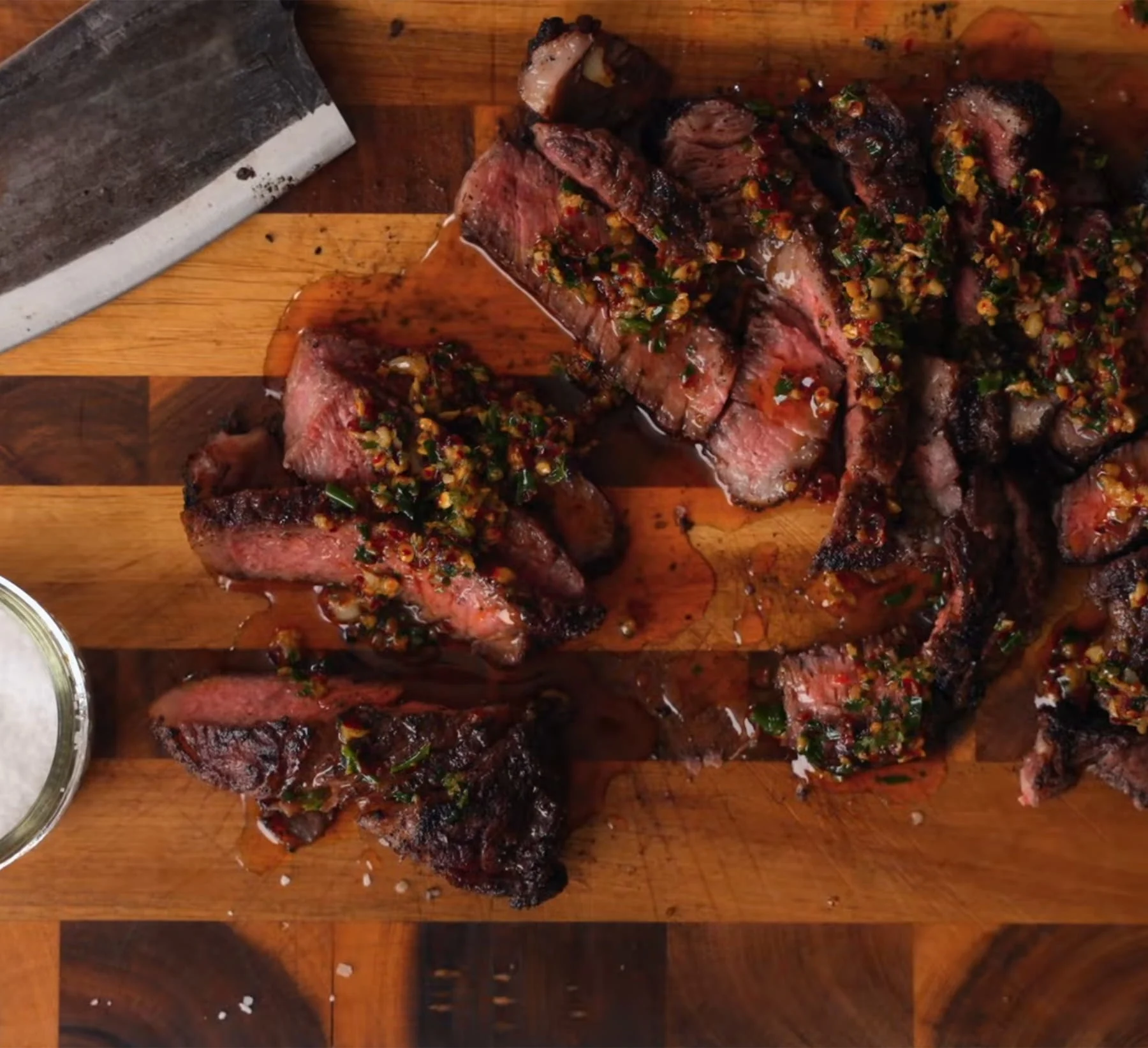 Slices of seared steak on a wooden cutting board with a knife and a ramekin of salt
