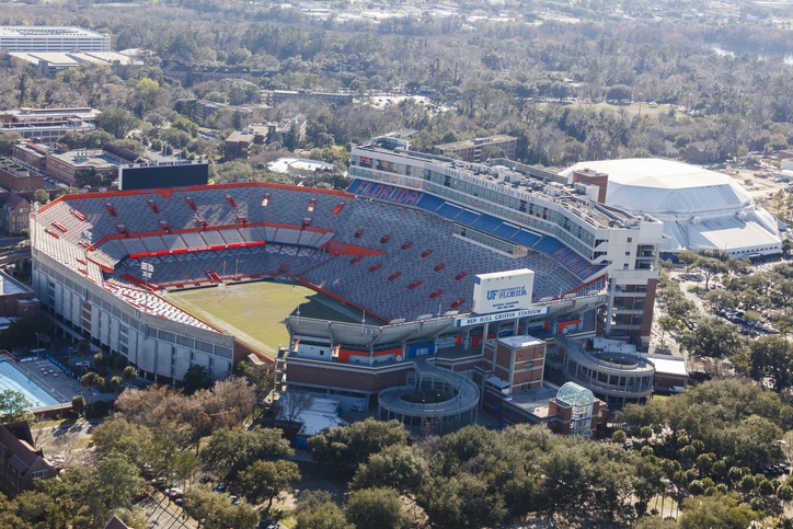 Aerial view of an empty Ben Hill Griffin Stadium in Gainesville, with various buildings and many more tall green trees nearby