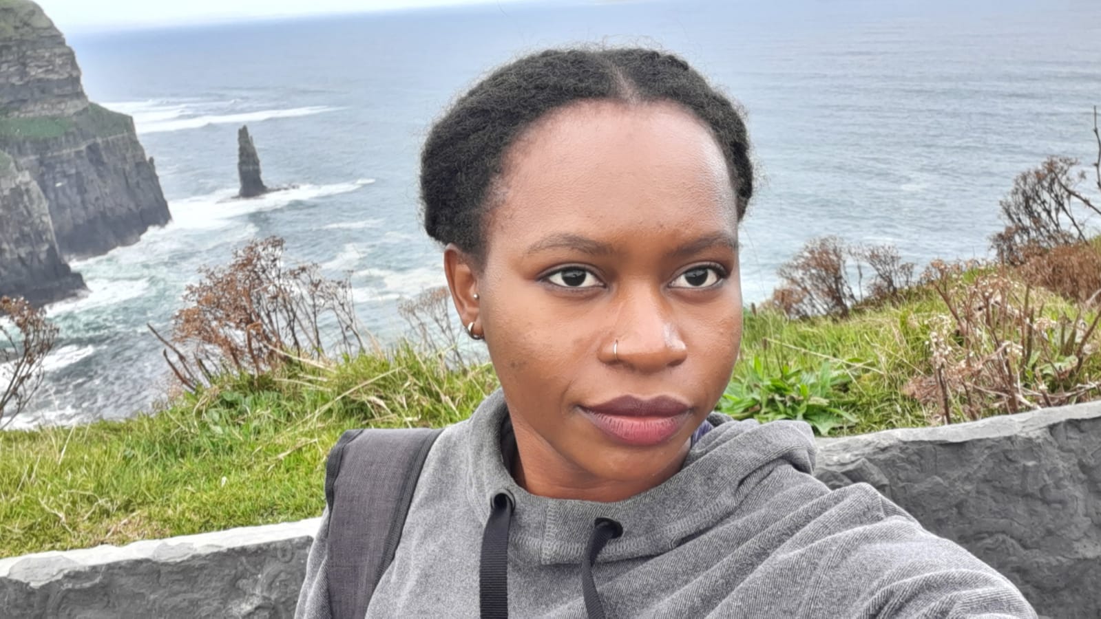 Sarah Lavender Achieng stands atop the Cliffs of Moher in Ireland, with rugged rocks and the Atlantic Ocean below and behind her