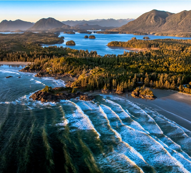 Aerial vie of A beach in Tofino, Vancouver Island in Canada.You can see white waves in the foreground lapping on shore with a series of island with hills and trees in the background.