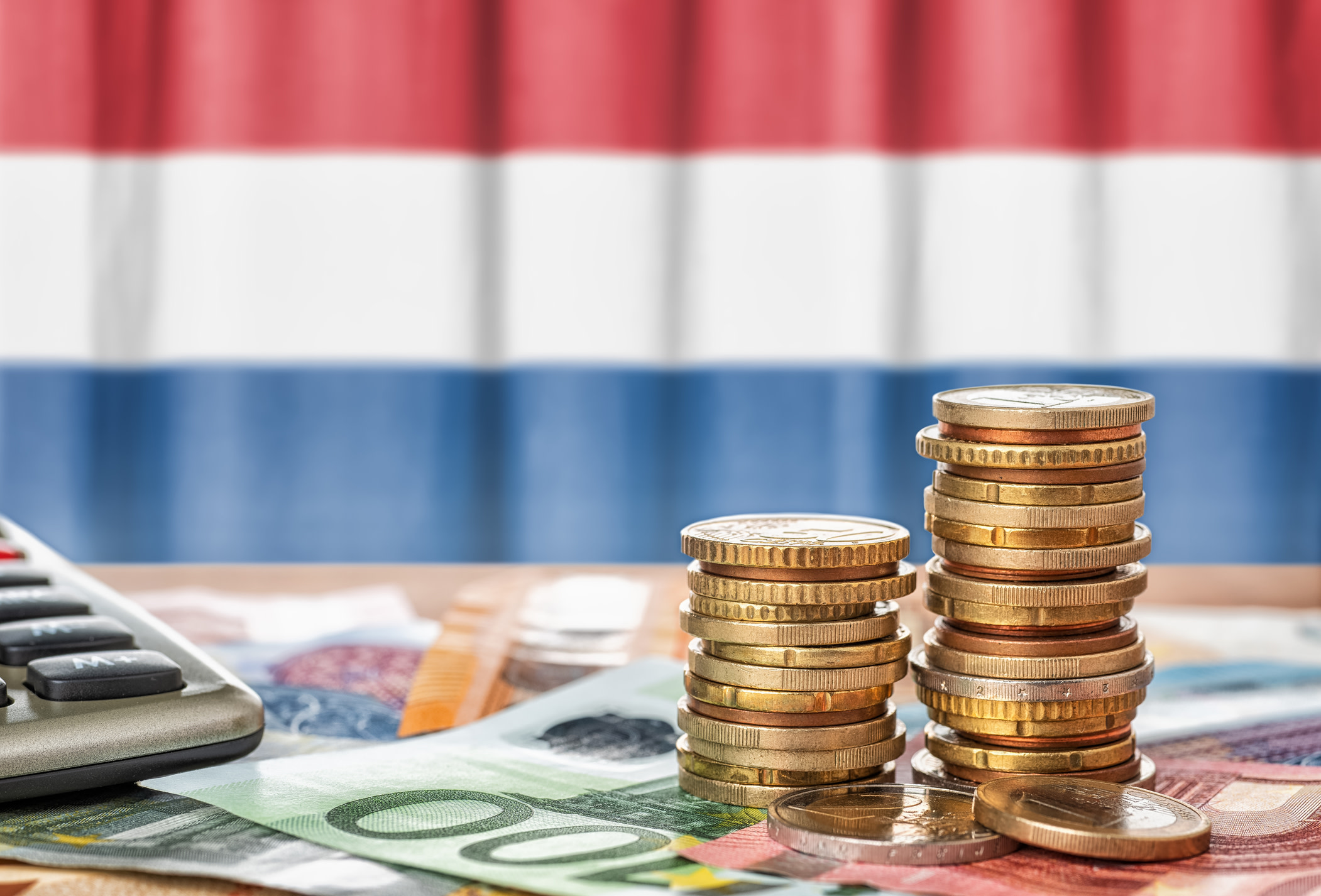 Graphic representing Netherlands tuition fees, with many Euro coins and notes placed on a table with a calculator in front of the Dutch flag