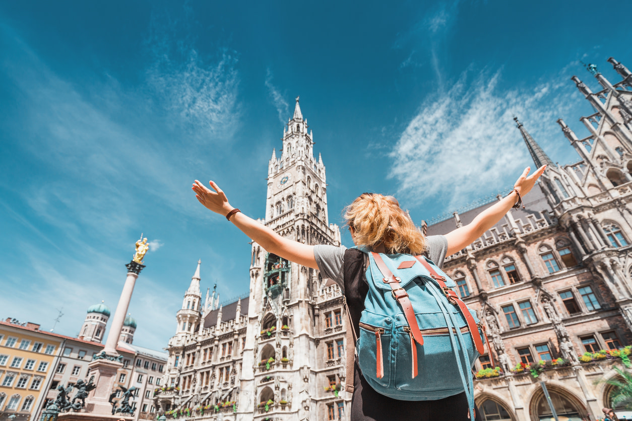 A young woman wearing a backpack raises her arms jubilantly in front of the New Town Hall in Munich on a sunny day