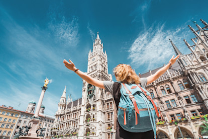 A young woman wearing a backpack raises her arms jubilantly in front of the New Town Hall in Munich on a sunny day