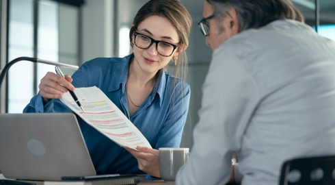 An investment banker shows her client some details on a piece of paper while using a laptop.