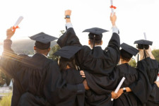 Group of students at graduation. The photo is taken from behind. The students are wearing graduation robes and caps, and holding their degrees