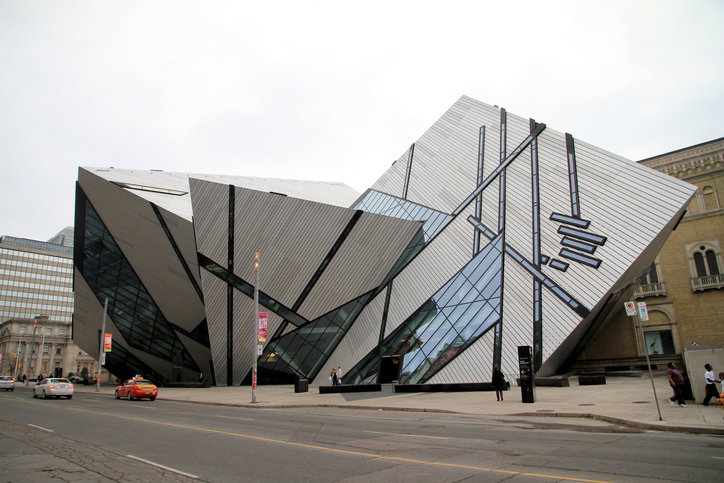 The jagged, three-peaked frontage and entrance of the Royal Ontario Museum. Several people and three cars are outside it.