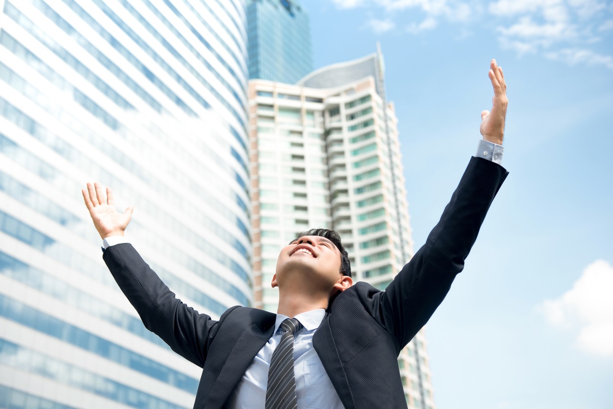 Young man in suit smiles broadly as he raise his arms and head up to a cloudy blue sky with tall glass buildings behind him