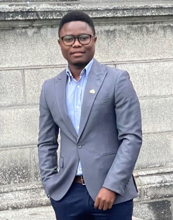 Emmanuel Sandy smiles in front of a grey concrete wall while wearing black glasses, a navy suit jacket, light blue shirt and black trousers.