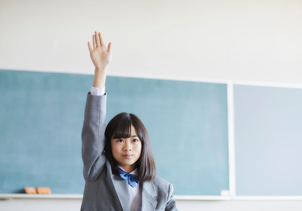 Seorang pelajar perempuan berseragam sekolah yang sedang mengangkat tangan untuk meminta bantuan dari pengawas tes IELTS