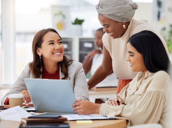 Three smiling women - two sitting, one stood - are by a laptop on a table that has various note papers and a coffee cup on it. Another woman sits behind them.