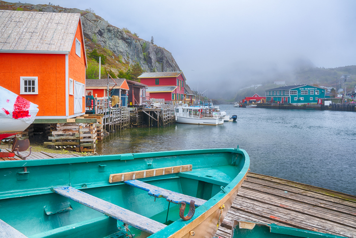 A small green-and-brown boat stands on a wooden jetty next to the water beside wooden buildings and a small cliffside in St John’s. Mist covers the water just ahead.