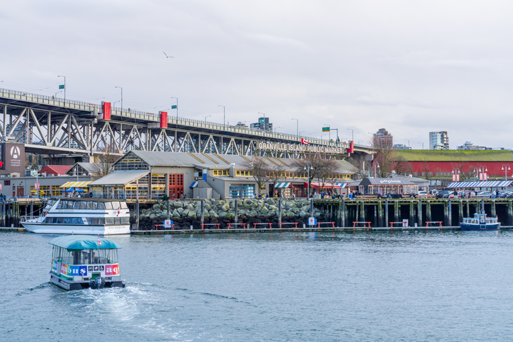 Granville Island Public Market. Vancouver marina. Vancouver, Canada. Standing under a bridge to the left and on the waterfront. There are two boats on the river. One is docked by the market the other is heading in that direction. 