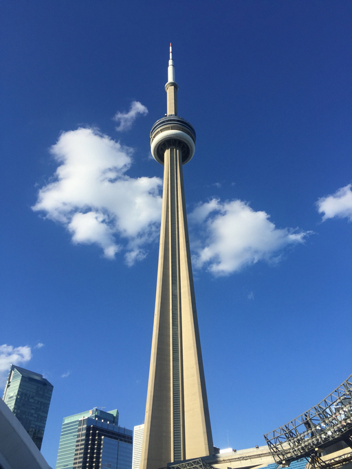 The thin and pointed CN Tower reaches high into the near-cloudless blue sky over Toronto; two other high-rise buildings are behind it.