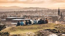 Thirteen young adult friends sit on the side of Arthur's Seat, an extinct volcano in Edinburgh. They have a view out across the city's skyline at dusk.