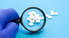 A hand covered in protective gloves holds a magnifying glass over a pile of circular white pills