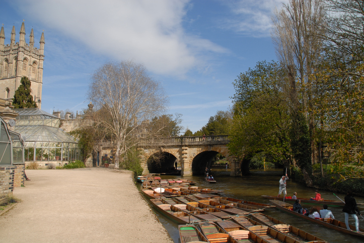 Several wooden punt boats rest on the water beside a smooth path, low bridge and tall trees in Oxford. A historic building is to their left.