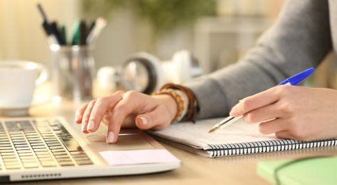 A student makes notes in a notebook while using a laptop computer