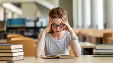 A stressed student in a library, looking at a book and holding her head