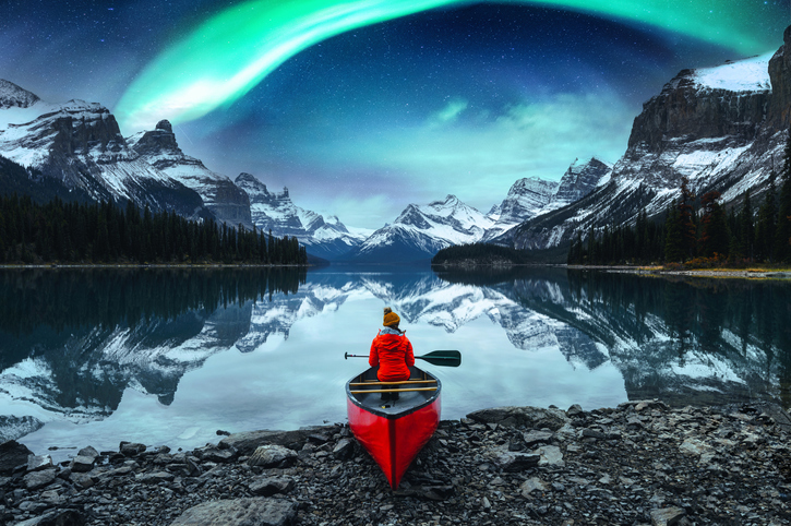 A woman sits in a red canoe by Maligne Lake in Jasper National Park, Alberta, as the Northern Lights shine over the mountainous, snow-capped Spirit Island ahead of her.