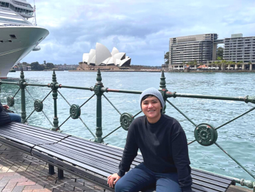 Joey sits smiling on a bench in front of Sydney Harbour and Sydney Opera House
