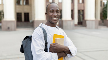 black male student with backpack and books posing over university building