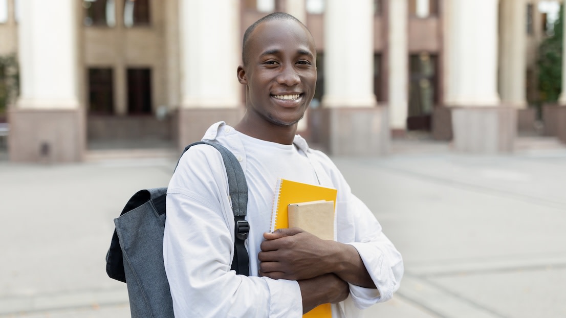 black male student with backpack and books posing over university building
