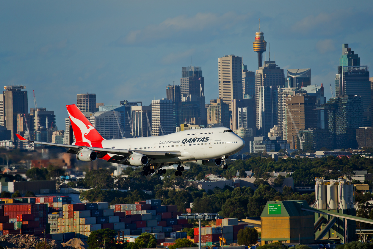 A Qantas plane flies low as it approaches landing, with many tall buildings filling the Sydney skyline behind it