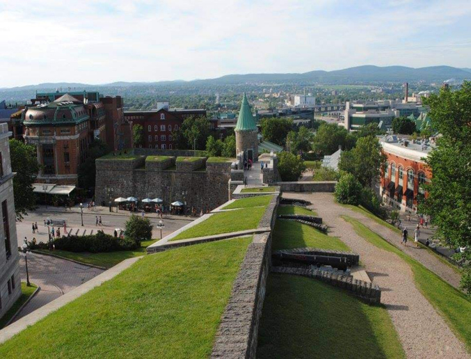 Many people walk near the fortified walls of Old Quebec City, surrounded by several historic buildings including the Saint-Louis Gate. Hills stand in the distance.