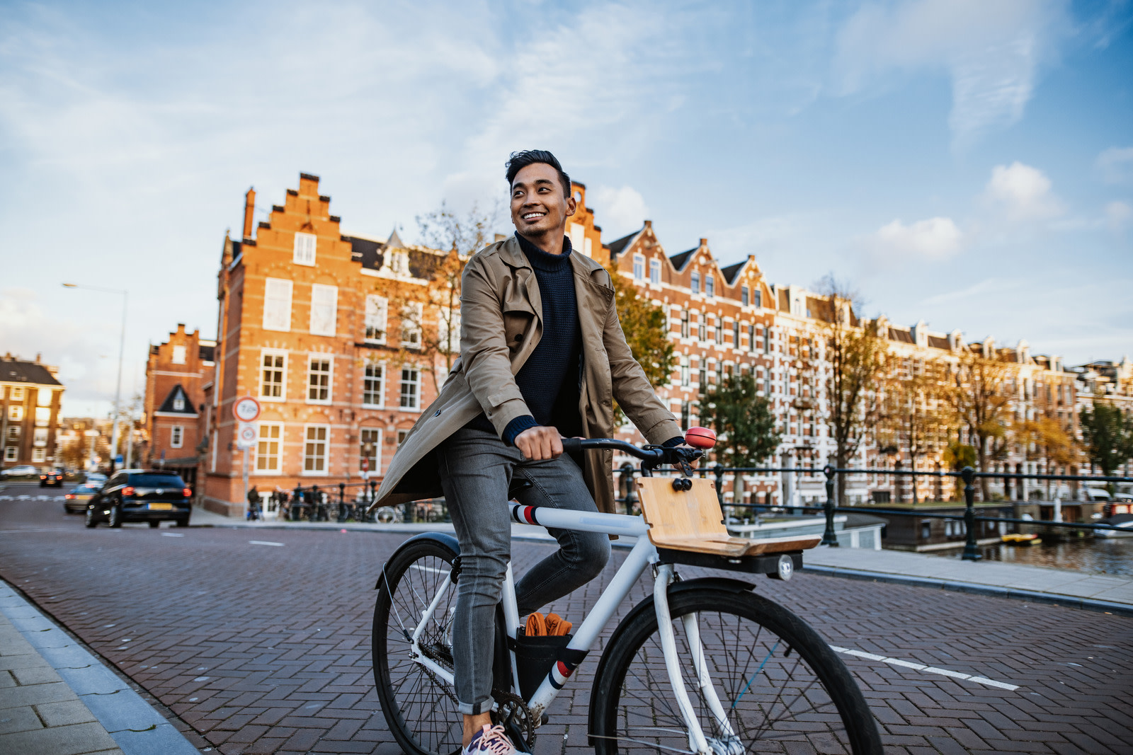 An international student smiles as he cycles across a bridge over a canal in Amsterdam, with the city's iconic canalside buildings behind him