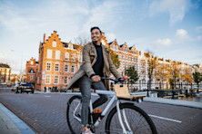 An international student smiles as he cycles across a bridge over a canal in Amsterdam, with the city's iconic canalside buildings behind him