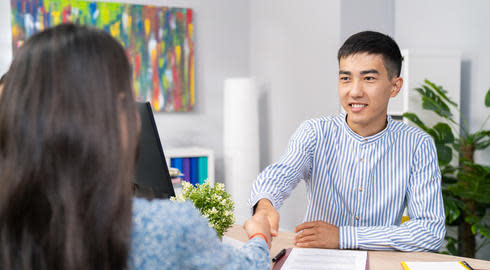 International student sitting at a desk. He is smiling at the person sitting in front of him. The person has her back to the camera. 