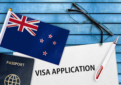 A visa application form rests on a blue wooden table alongside a pen, passport, pair of glasses and flag of New Zealand