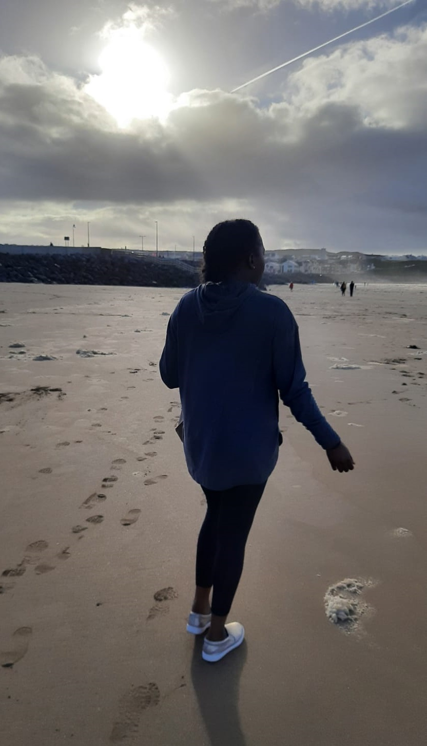 Sarah Lavender viewed from behind on a wet sandy beach, looking towards a cloudy sky through which the sun is breaking