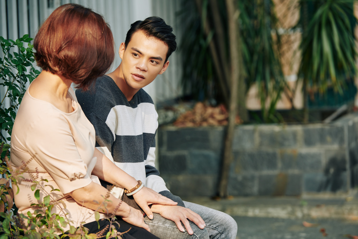 Young man sits near a wall with a palm tree on it. He looks thoughtfully at an older woman who has her hand placed on top of his.