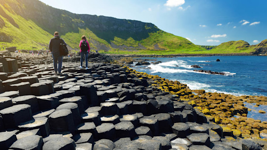 People walking on the Giants Causeway, on an area of hexagonal basalt stones, by green hills, blue skies and blue sea