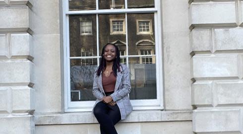 Lebokai sitting on a window sill outside a large university building. She is wearing black tights, black skirt, a brown top and a black and white checked jacket. She is smiling warmly.