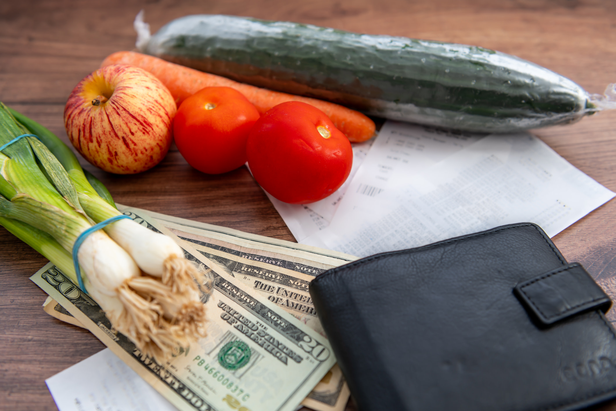 A pile of US Dollar bills, grocery receipts, a wallet and some vegetable groceries on a table.