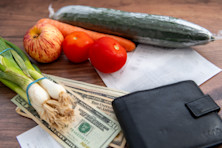 A pile of US Dollar bills, grocery receipts, a wallet and some vegetable groceries on a table.