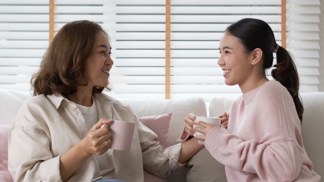 A mother and daughter smile while holding a mug each as they sit on a white sofa in front of white horizontal blinds.