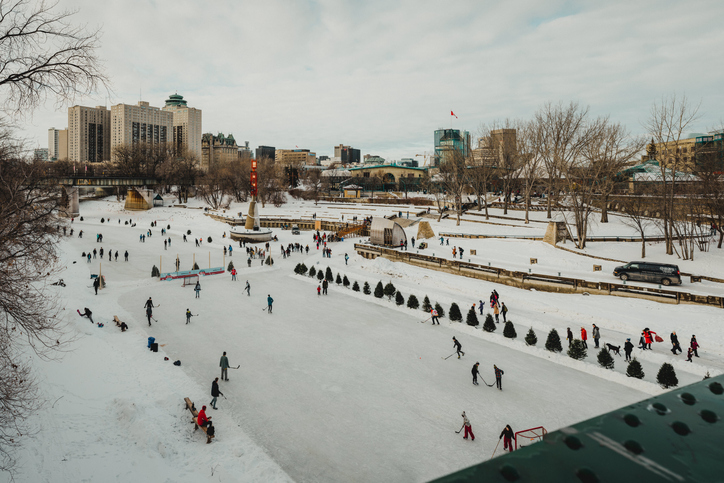 Many people play ice hockey and skate across frozen rivers in Winnipeg. In the background are several of the buildings that make up The Forks recreation district.