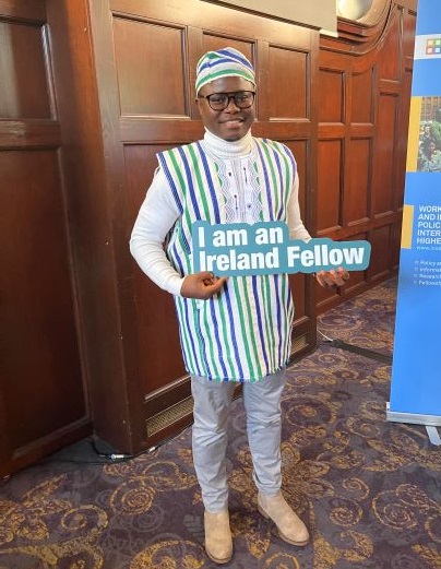 Emmanuel Sandy smiles while holding a sign reading 'I am an Ireland fellow' in a wood-panelled room with an elaborately designed carpet.